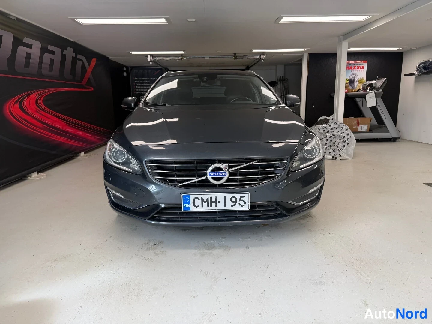 Edestä: Tummanharmaa Volvo V60 etukädestä kuvattuna huoltamotilassa, rekisterikilpi.