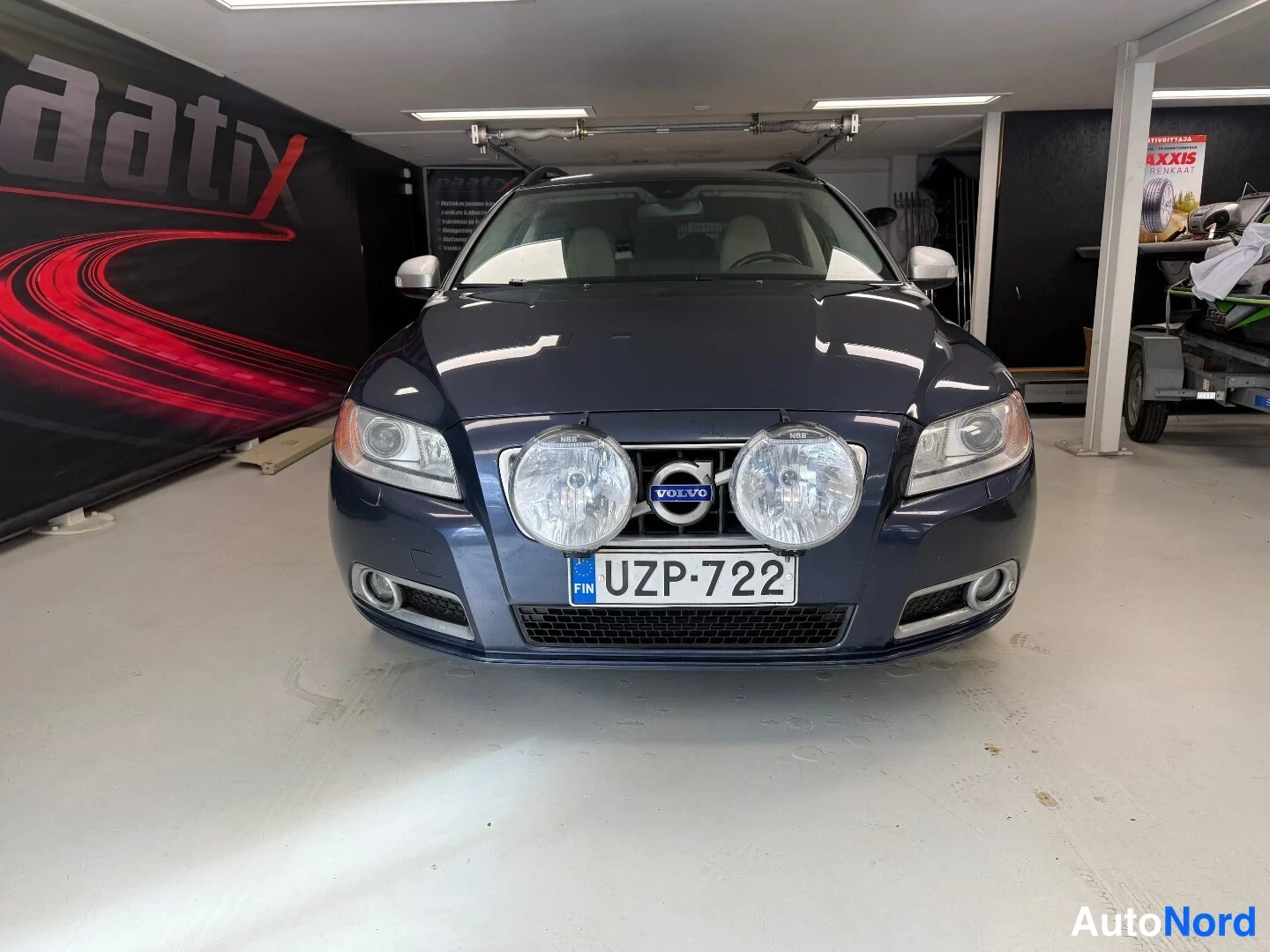 Edestä: Volvo V70 -farmarin etupuoli sinisenä, keulassa kaksi lisävaloa ja -kilpi sisätiloissa.