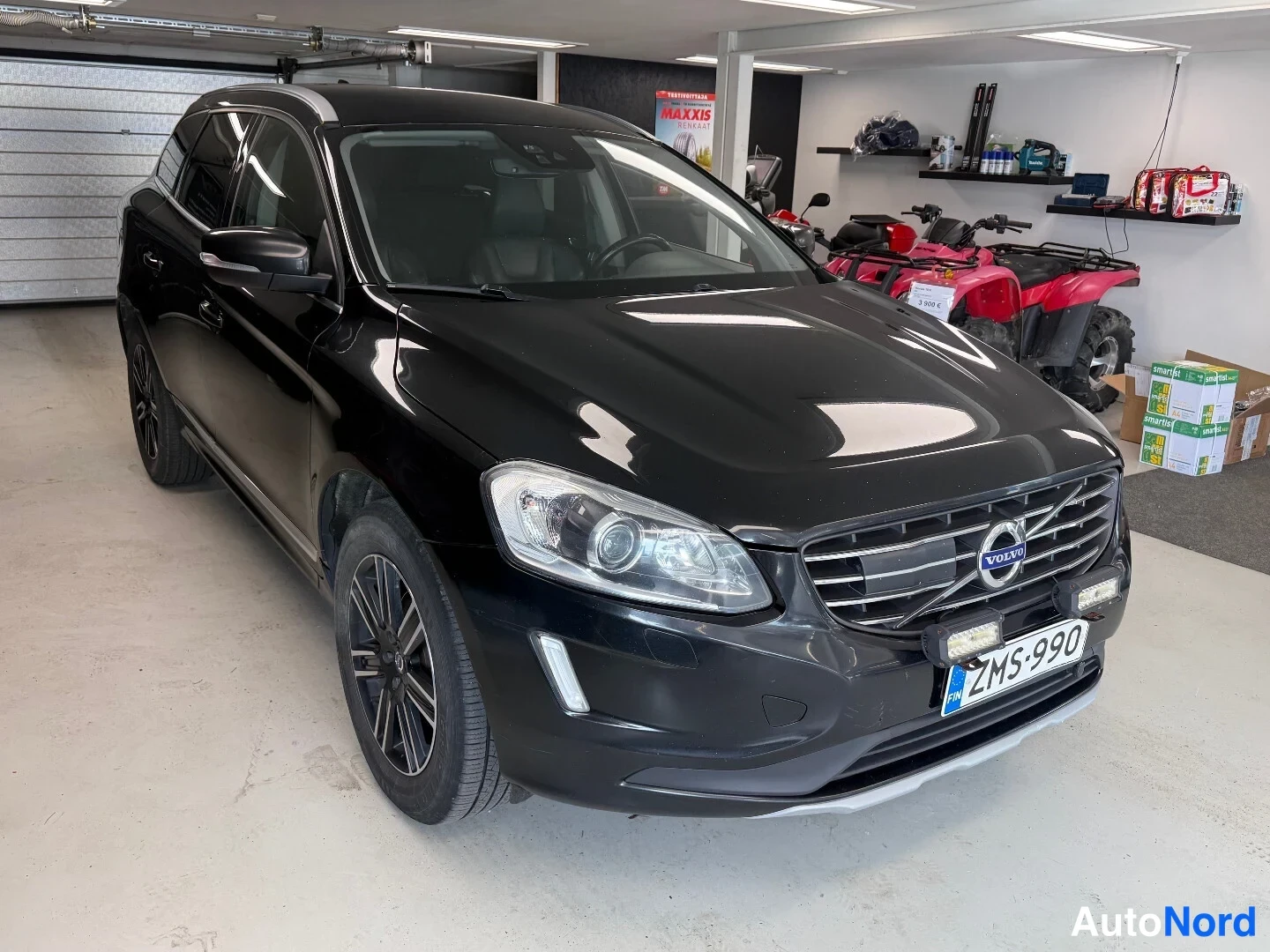 Etuviisto, nokka oikealle: Musta Volvo XC60 etukulmassa vasemmalta kuvattuna autotallissa, Volvo-merkki säleikössä ja lisävalot puskurissa näkyvät.