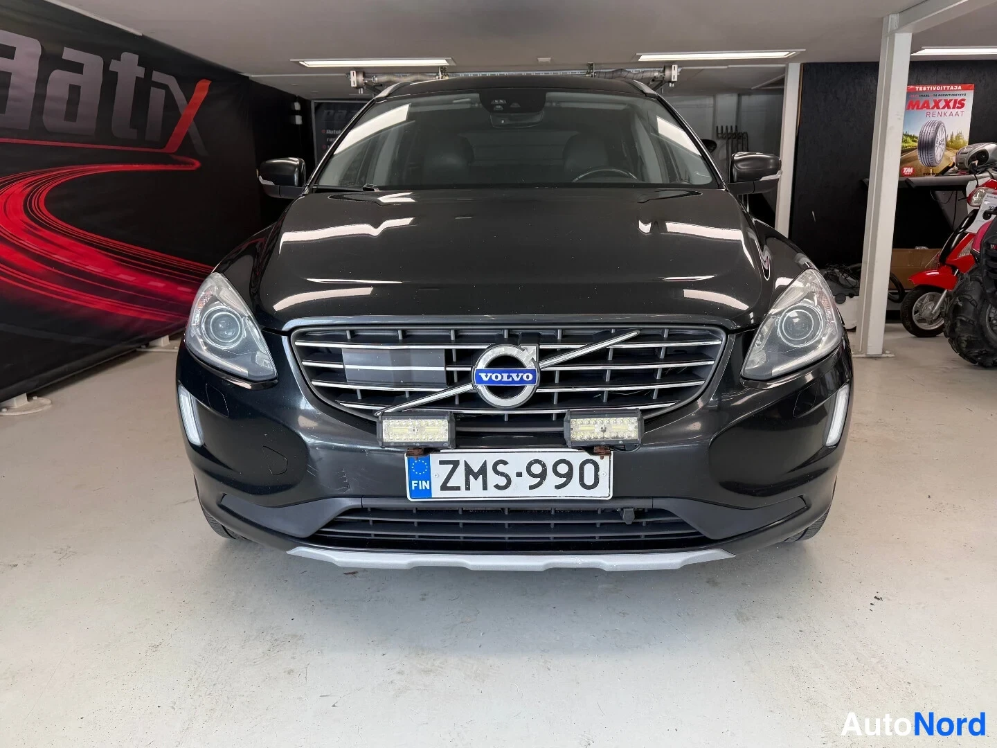 Edestä: Mustan Volvo XC60:n etupää, Volvo-merkki ja rekisterikilpi, lisävalot puskurissa.