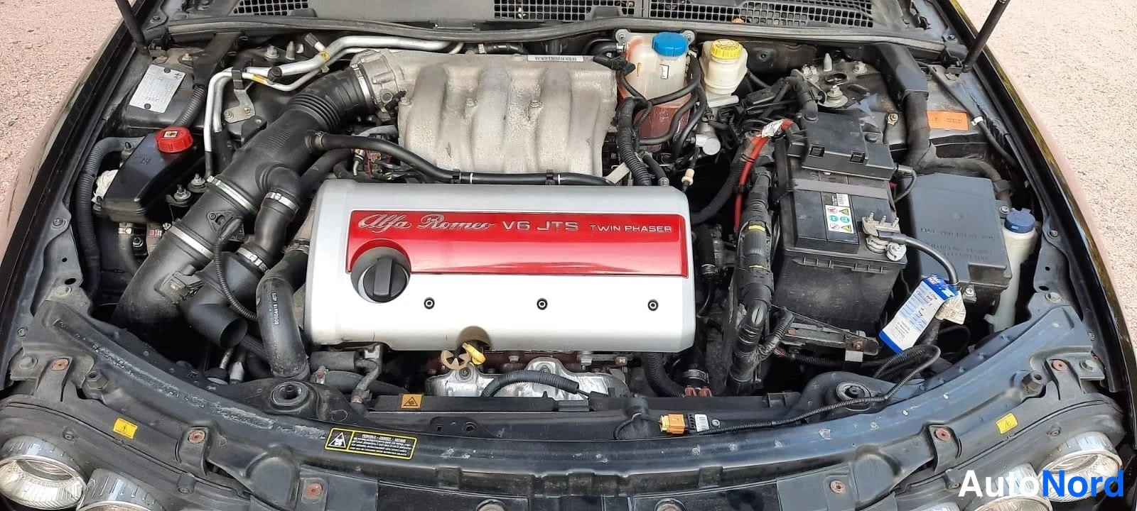 Moottoritila: Moottoritilan ylänäkymä: Alfa Romeo V6 JTS Twin Phaser -moottori, punakantinen sylinterinkansi, akku oikealla ja jäähdytysnestesäiliö sinisellä kannella.