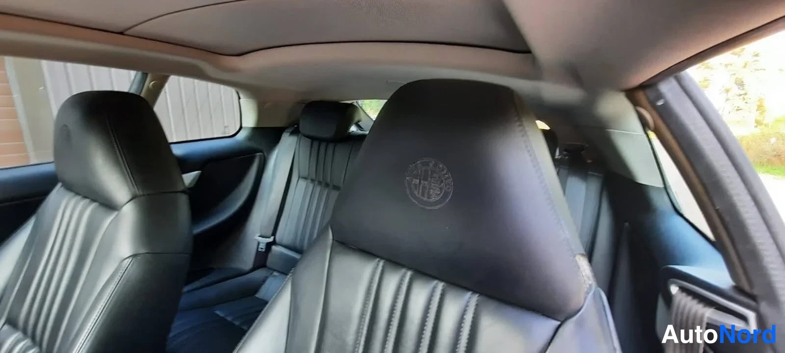 Muu: Auton takapenkin näkymä: mustat nahkaistuimet, etuistuinten selkänojissa Alfa Romeo -logot ja lasikattoaattana.