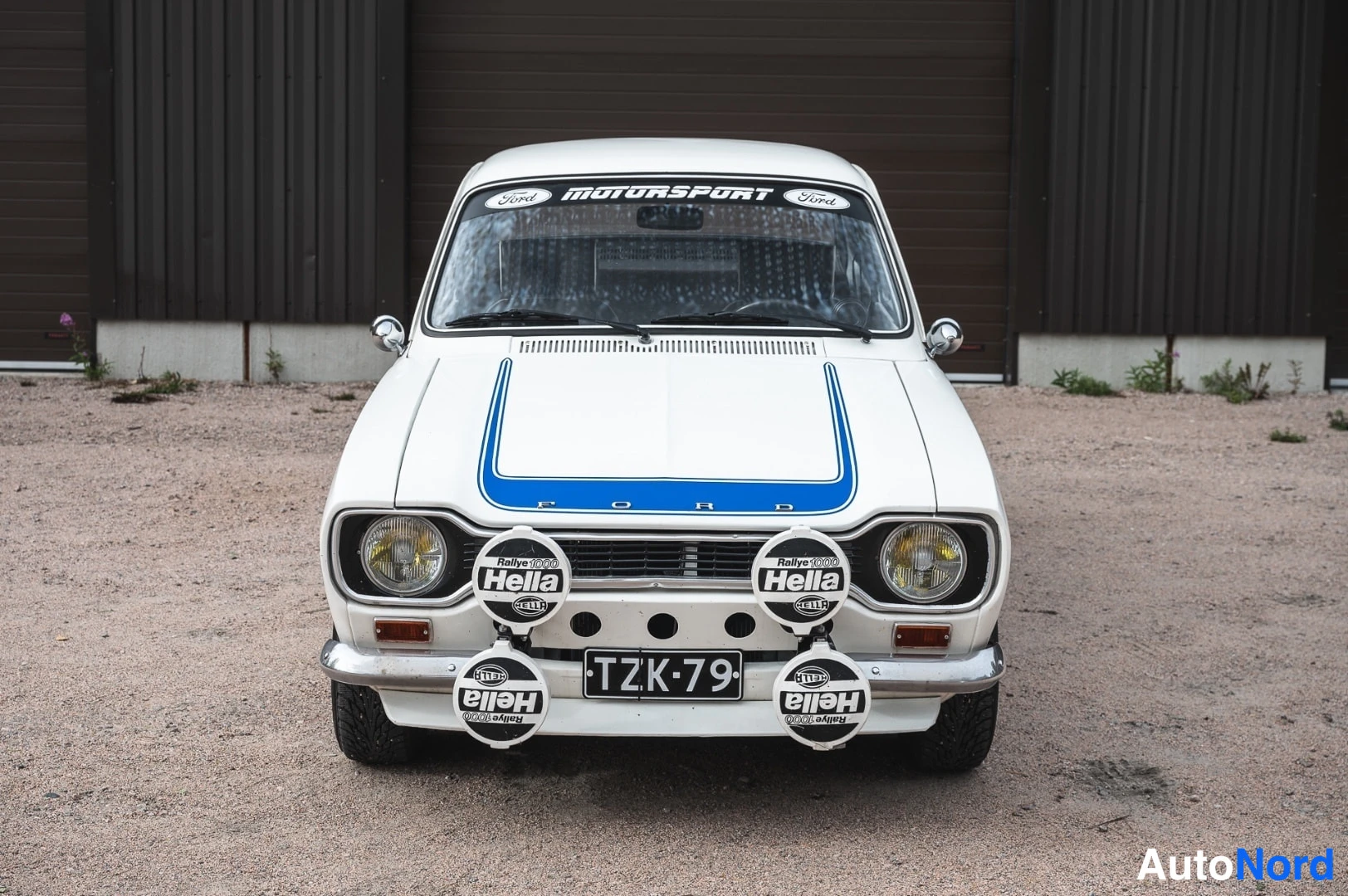 Edestä: Valkoinen klassinen Ford Escort suoraan edestä, konepellillä sininen koriste ja 'FORD'-teksti, neljä Hella-lisävaloa edessä.