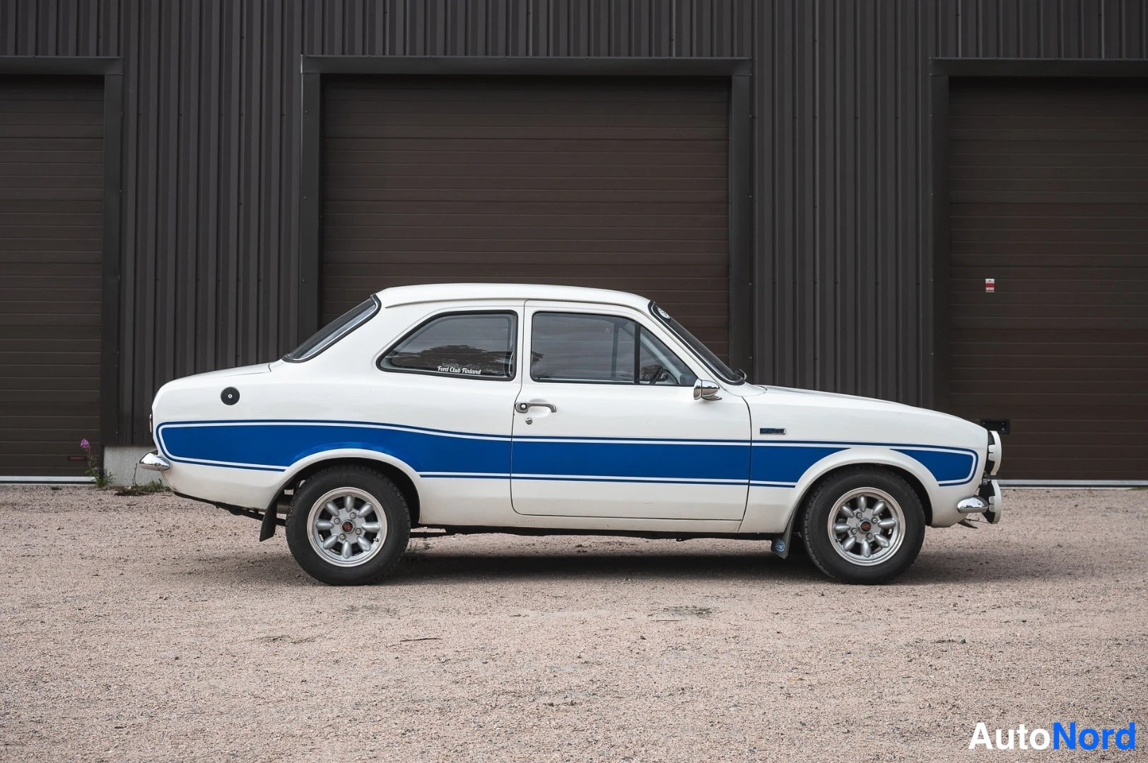 Etuviisto, nokka oikealle: Valkoinen Ford Escort sinisellä sivukaistaleella, kolmikulmakuva edestä oikealta.