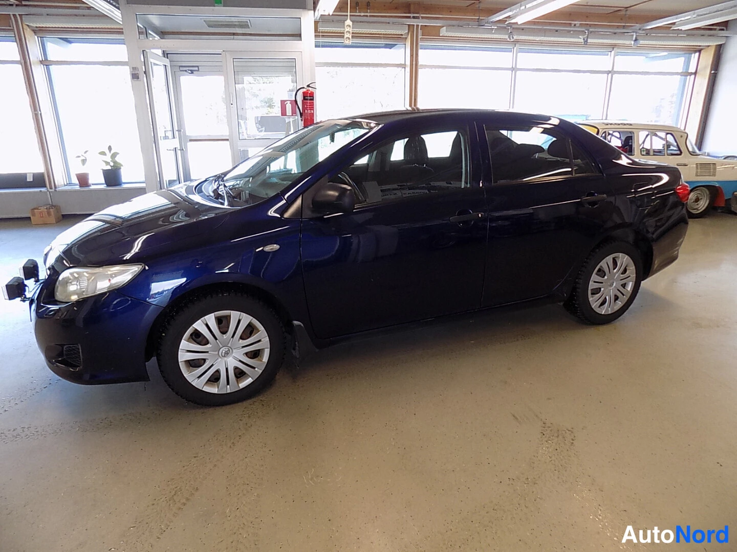 Etuviisto, nokka vasemmalle: Tummansininen Toyota Corolla -sedan etunurkkakuva vasemmalta sisätilassa, vasemmat ovet ja etupuskuri näkyvät.