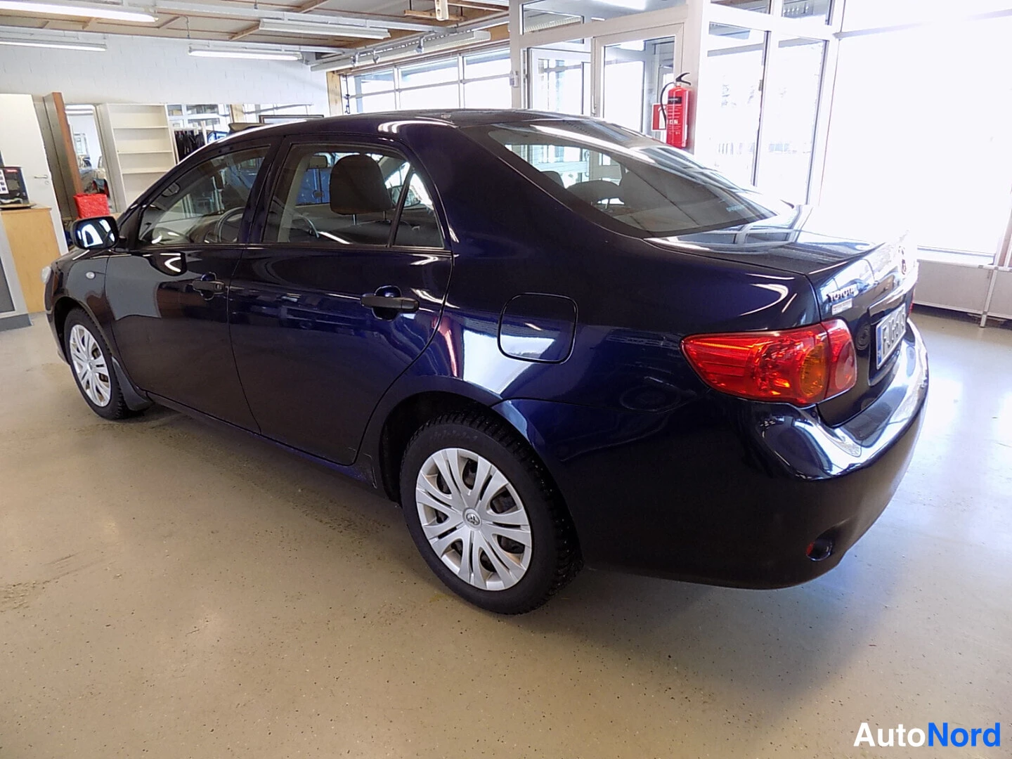 Takaviisto: Tummansininen Toyota Corolla -sedan takanurkkakuva vasemmalta sisätilassa, takavalot ja vasemmat ovet näkyvät.