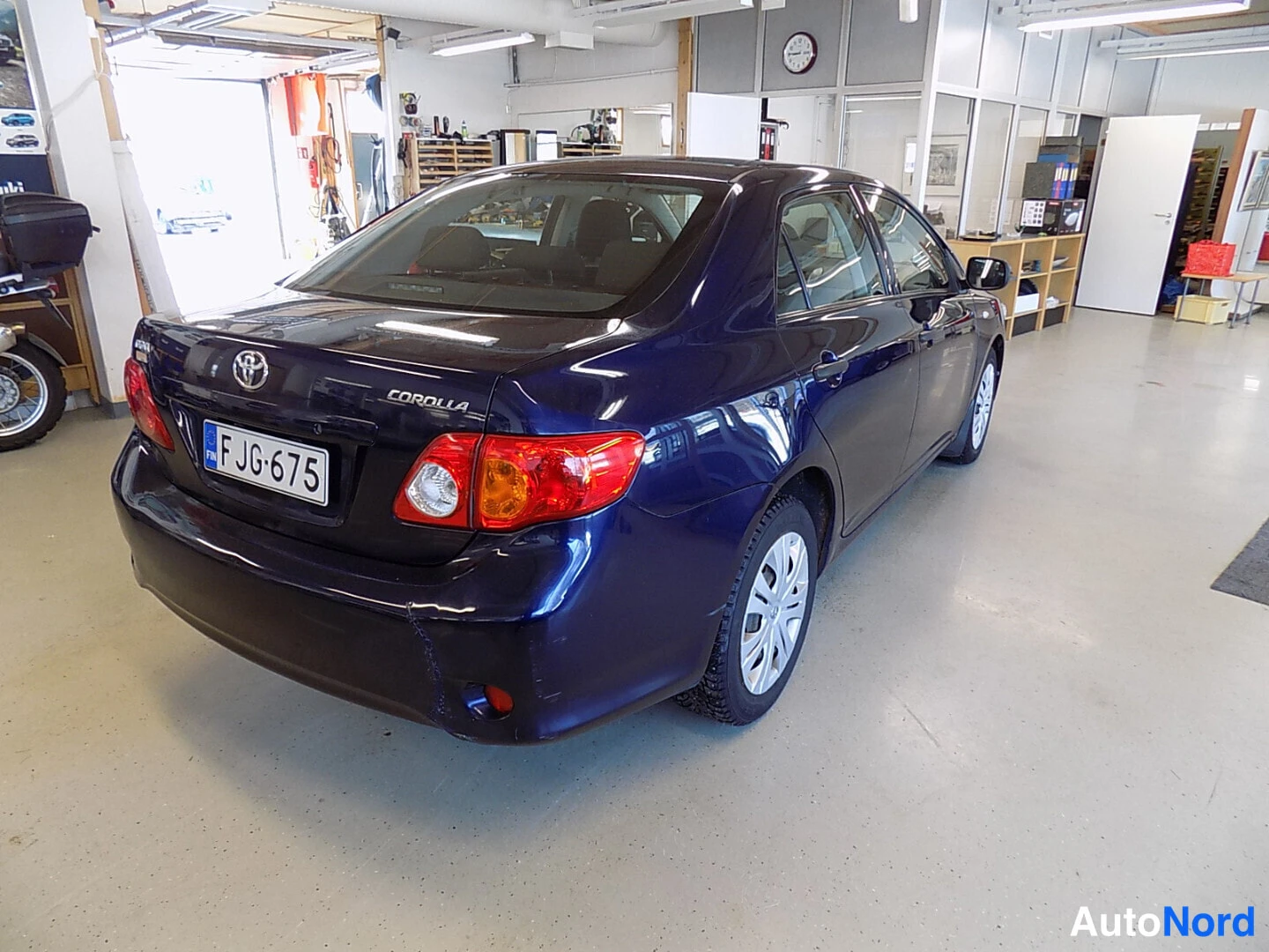 Takaviisto: Tummansininen Toyota Corolla -sedan takanäkymä sisätilassa, rekisteritunnus F JG-675 ja Corolla-merkintä takaluukussa näkyvät.