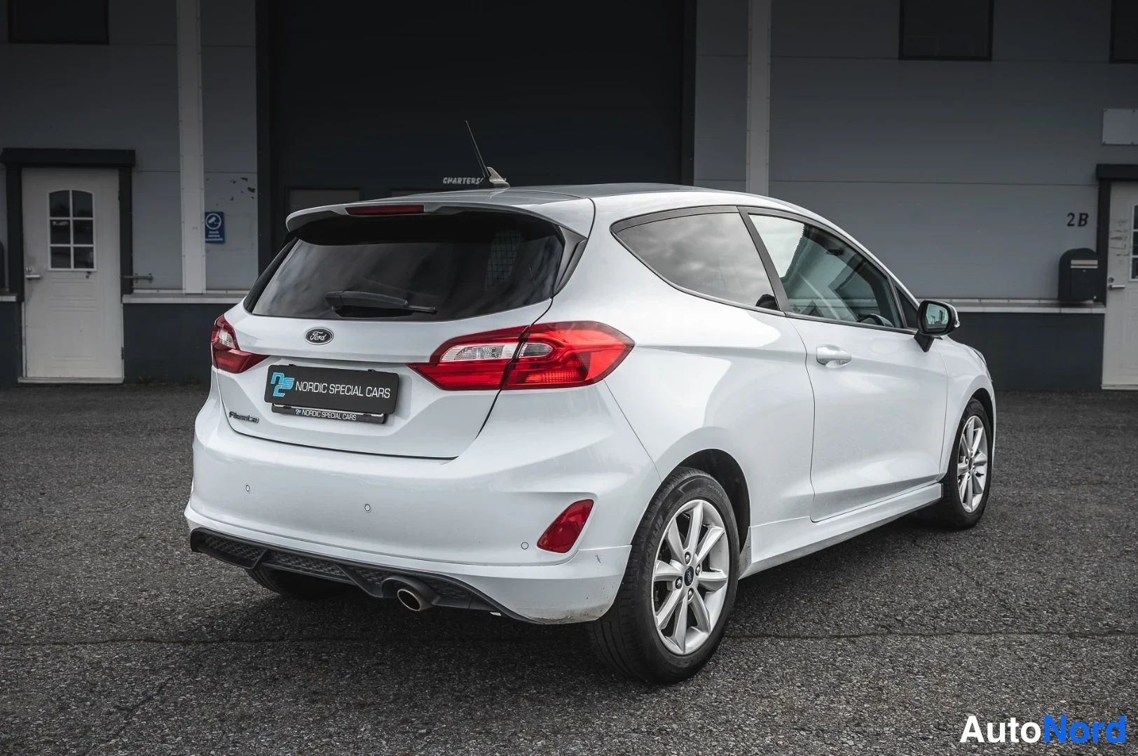 Takaviisto: Valkoinen Ford Fiesta taka-ultapäältä vasemmasta kulmasta, takavalot ja kaksiosainen pakoputki näkyvissä.