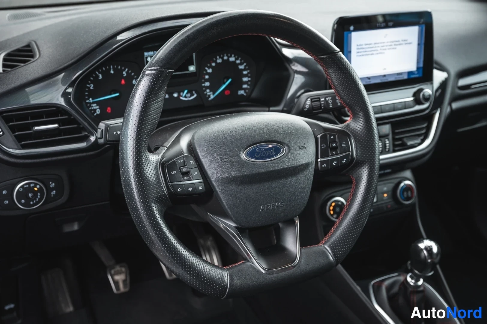 Takaa: Ohjauspyörän lähikuva Ford-merkillä, kojelaudan mittarit ja osittain näkyvä infotainment-näyttö.
