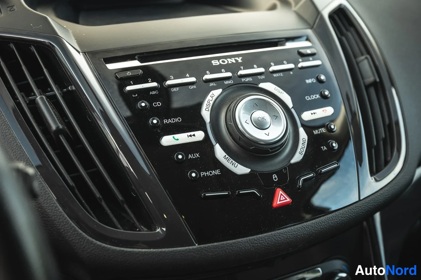 Yksityiskohta: Lähikuva Sony-autoradiosta: painikkeet CD, RADIO, AUX, PHONE, numeroitus 1–6, OK-ohjain ja varoitusvalon kolmiopainike.