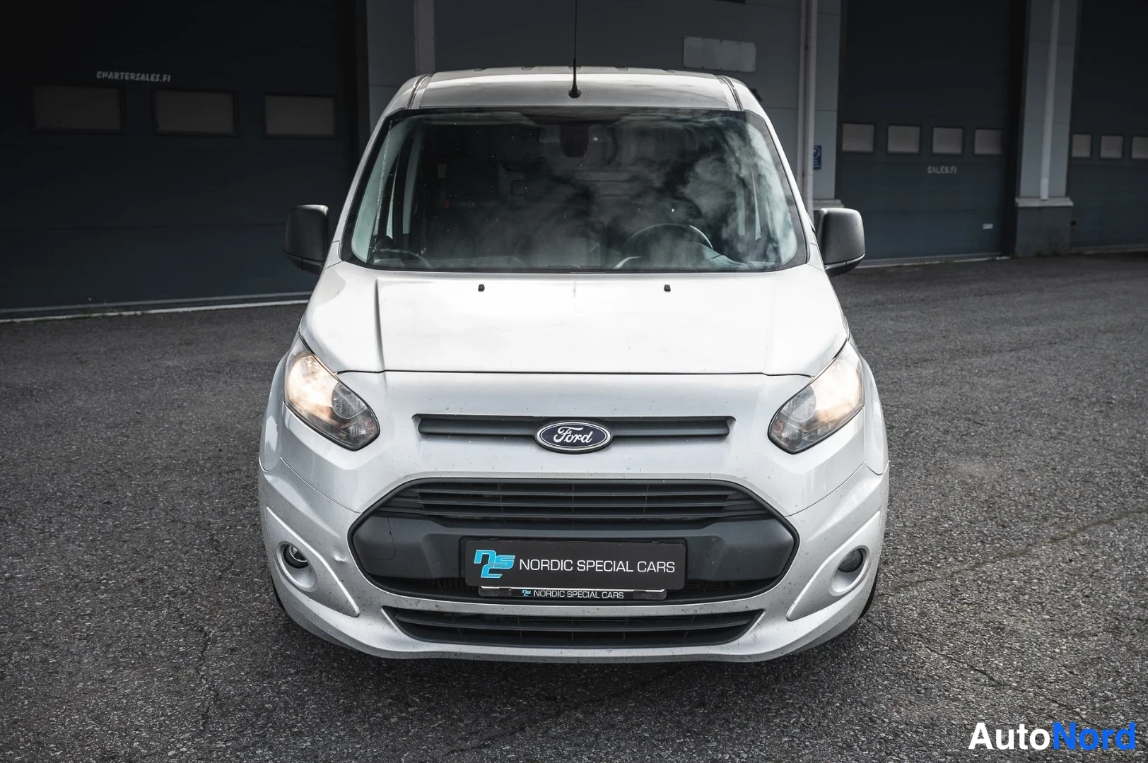 Edestä: Ford Transit Connect -pakettiauton etupää suoraan edestä valokuvattuna; hopeanharmaa kori, Ford-merkki etusäleikössä ja Nordic Special Cars -kyltti etupuskuriss