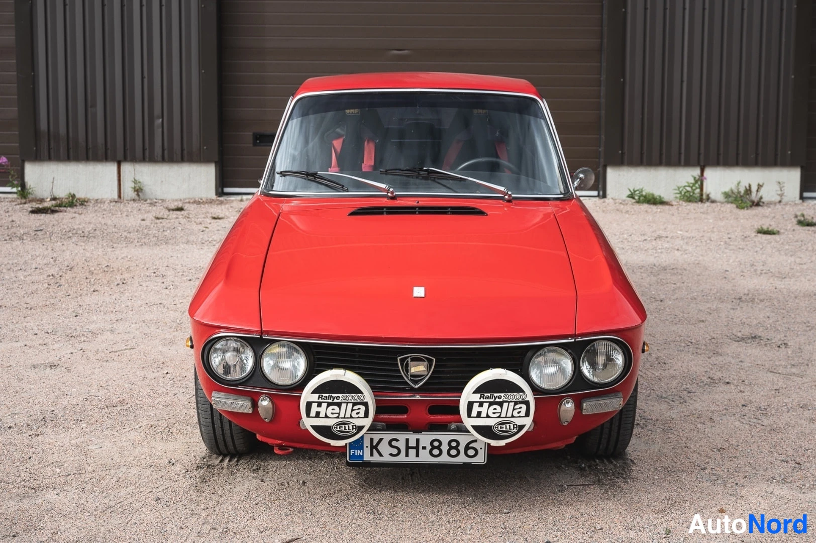 Edestä: Punainen Lancia Fulvia -coupe suoraan edestä, neljä pyöreää ajovaloa, kaksi Hella Rallye 3000 -lisävaloa ja rekisteri.