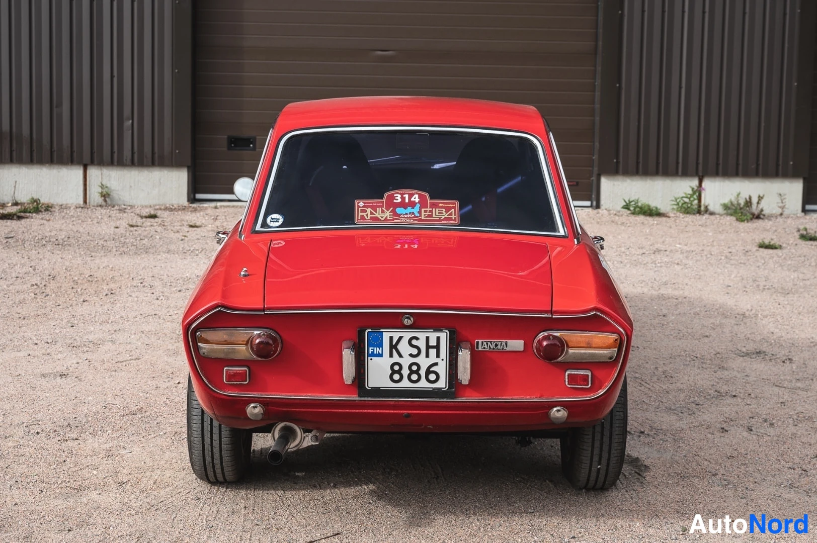 Takaa: Punainen Lancia Fulvia -coupe suoraan takaa, rekisteri, LANCIA-teksti ja Rallye Elba -tarra takalasissa.