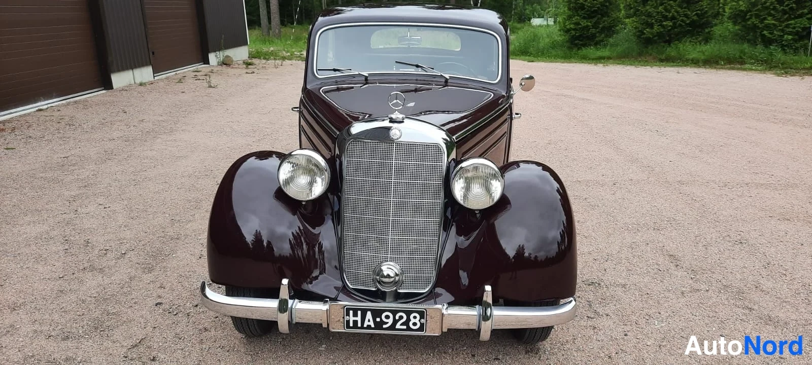 Edestä: Tummaviininpunainen Mercedes-sedan suoraan edestä, kromikehystetty jäähdyttingrilli, kaksi pyöreää ajovaloa ja rekisterikilpi HA·928.