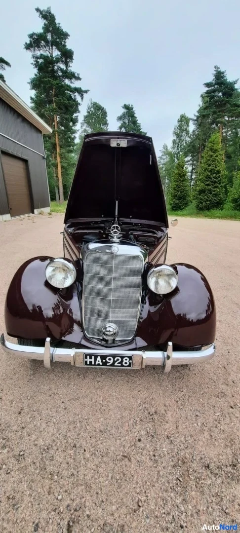 Edestä: Mercedes-Benzin tummanpunaisen klassisen auton etunäkymä: konepelti auki, korostettu jäähdytinsäleikkö ja kaksi pyöreää ajovaloa, etupuskurilla rekisteritunnus