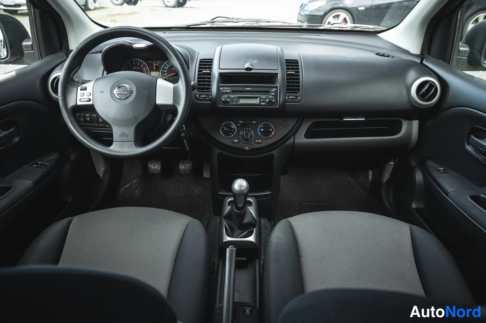 Muu: Etupenkki ja kojelauta edestäpäin kuvattuna; Nissan-ohjauspyörä, viisarit, radio, manuaali vaihteenvalitsin ja polkimet näkyvät.