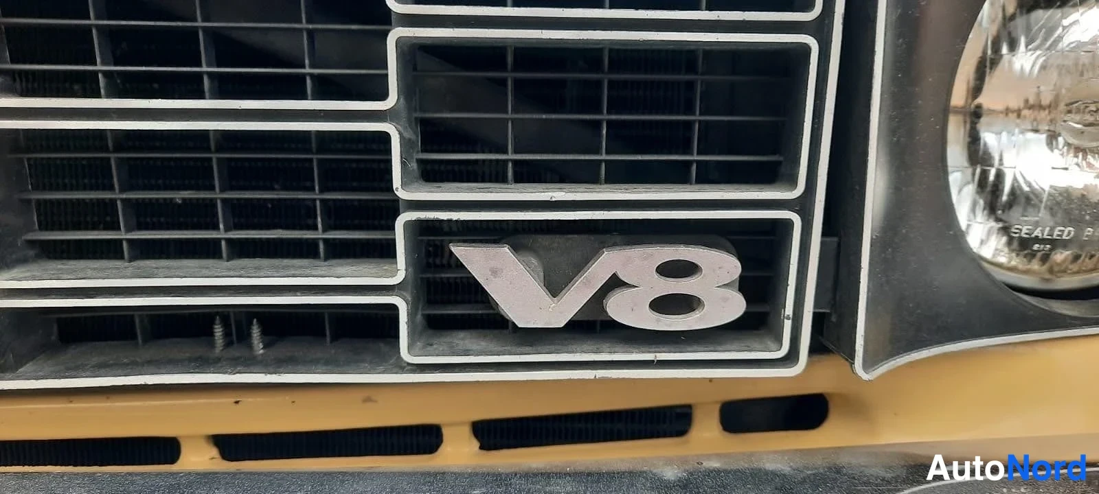 Edestä: Suljettu kuva auton etusäleiköstä, jossa V8-merkki ja osa pyöreästä ajovalosta.