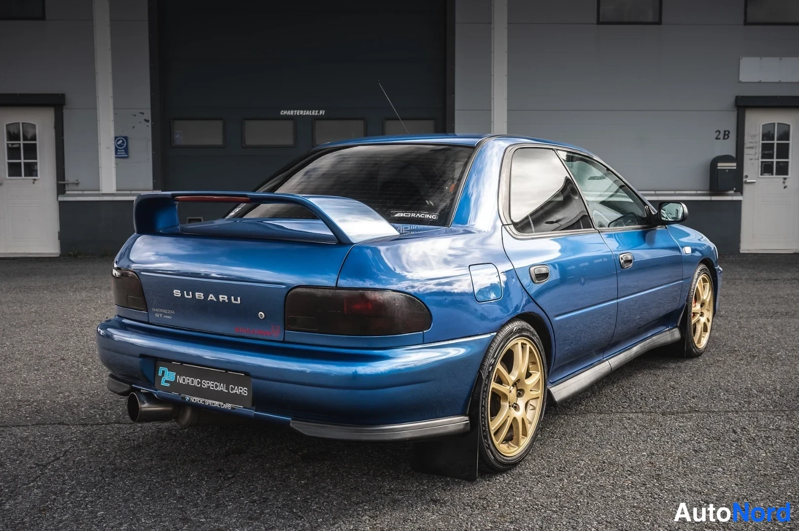 Takaviisto: Sinisen sedanin takakolmannes, SUBARU-teksti takaluukussa ja kultaiset vanteet.