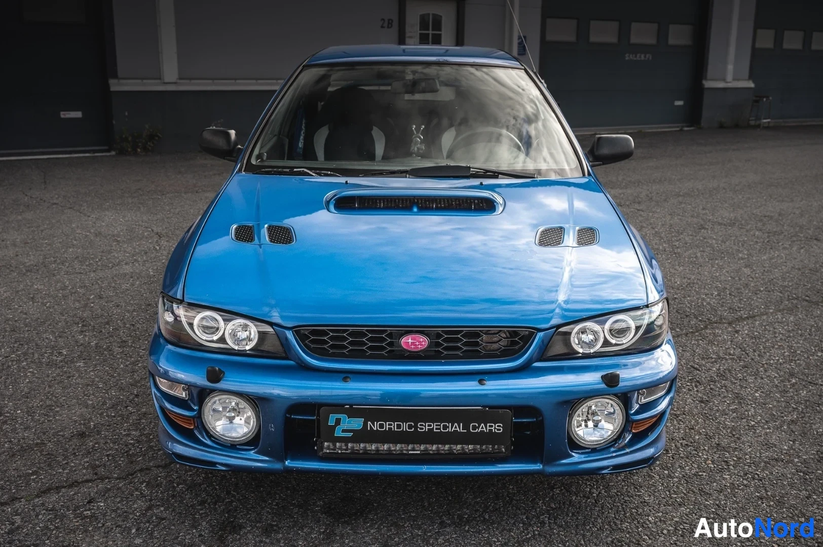 Edestä: Sininen Subaru Impreza edestä kuvattuna, syöttöaukko konepellissä ja kaksi lisävaloa puskurissa.