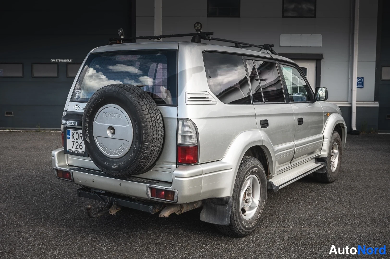 Takaviisto: Hopeisen Toyota Land Cruiser Pradon takaosa kolme neljännestä vasemmalta, varapyörä kannessa ja pyykinripustustanko perässä.