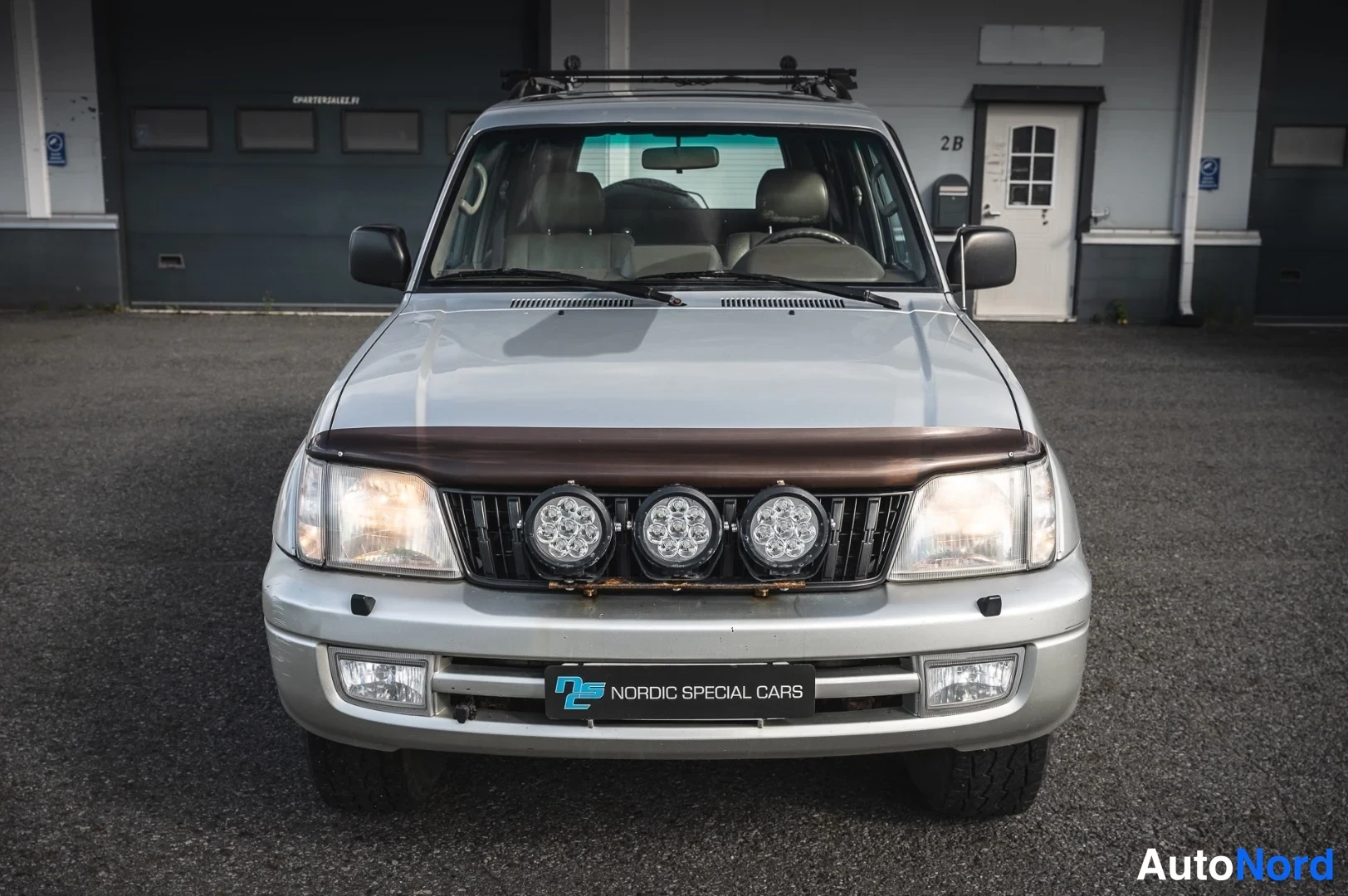 Edestä: Hopeanvärinen Toyota Land Cruiser Prado edestä kuvattuna; keulalla kolme lisävaloa ja kattoteline.