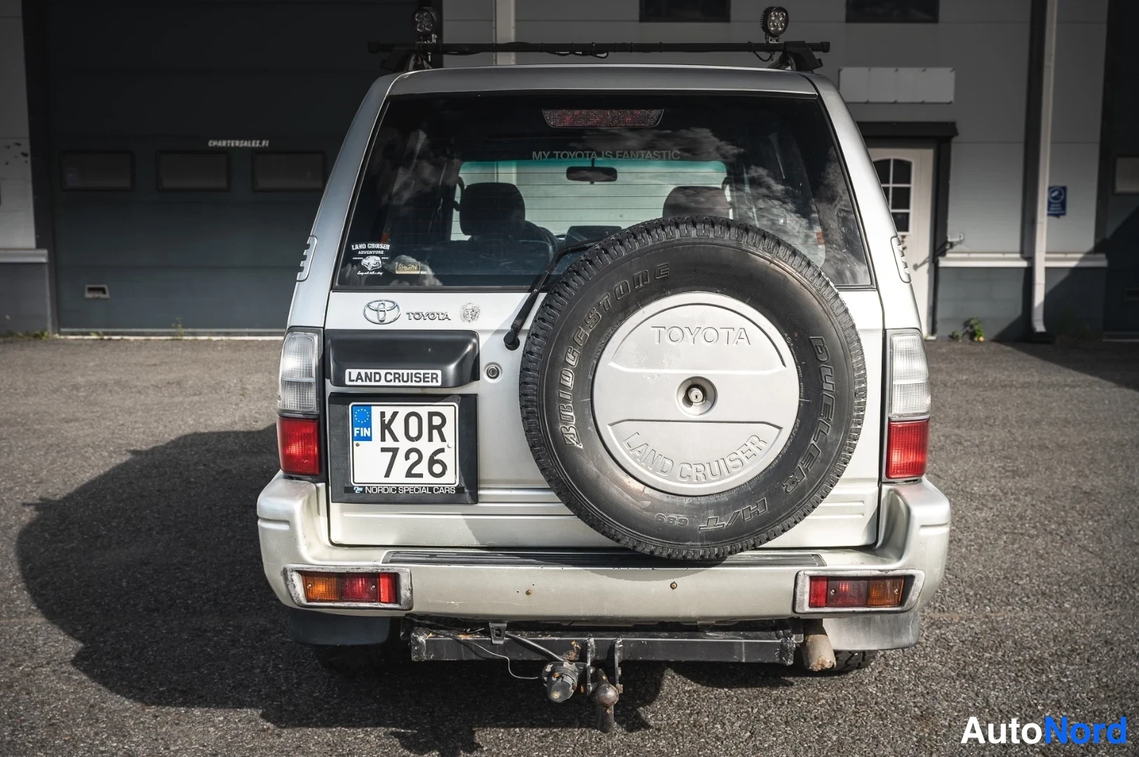 Takaa: Toyota Land Cruiserin takaosa: varapyörä TOYOTA-korilla, rekisterikilpi KOR 726, kattoteline ja vetokoukku.