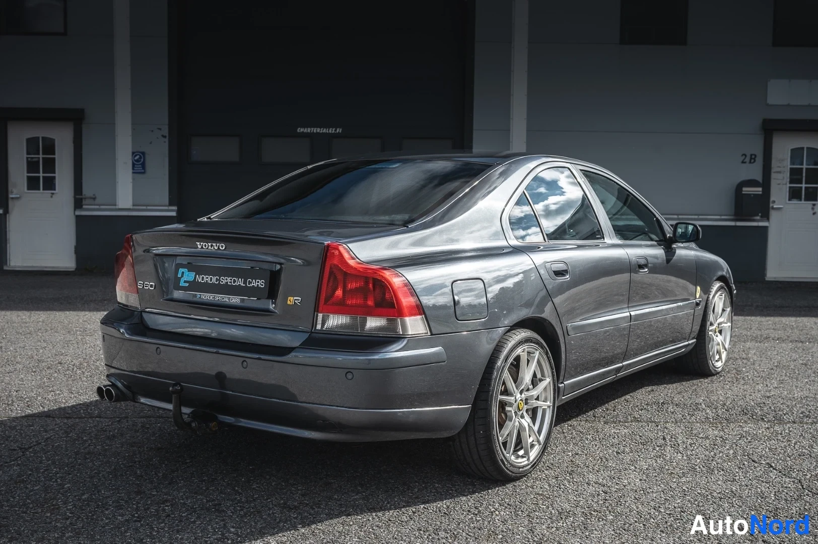 Takaviisto: Auton takakulmakuva: tummanharmaa S60-sedan, punaiset takavalot, kaksoispakoputket ja irrotettava vetokoukku.
