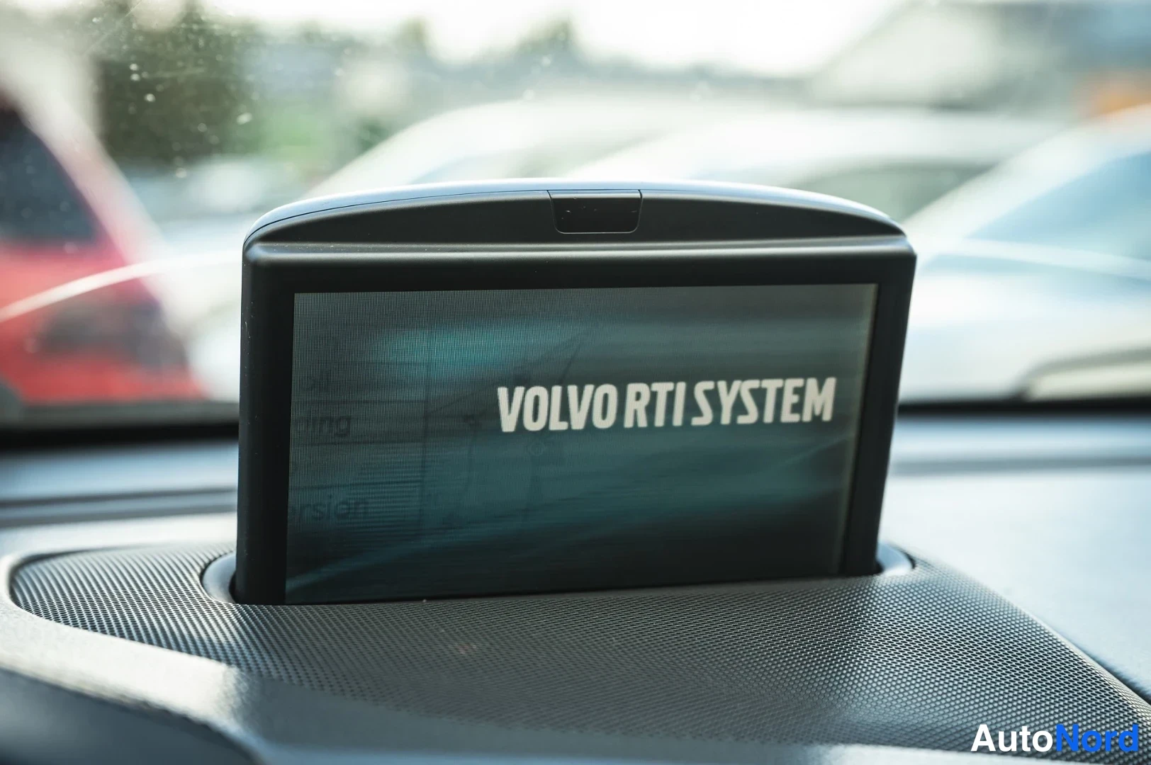 Muu: Kojelaudasta kääntyvä näyttö, jossa lukee VOLVO RTI SYSTEM; taustalla auton takana pysäköituja autoja.