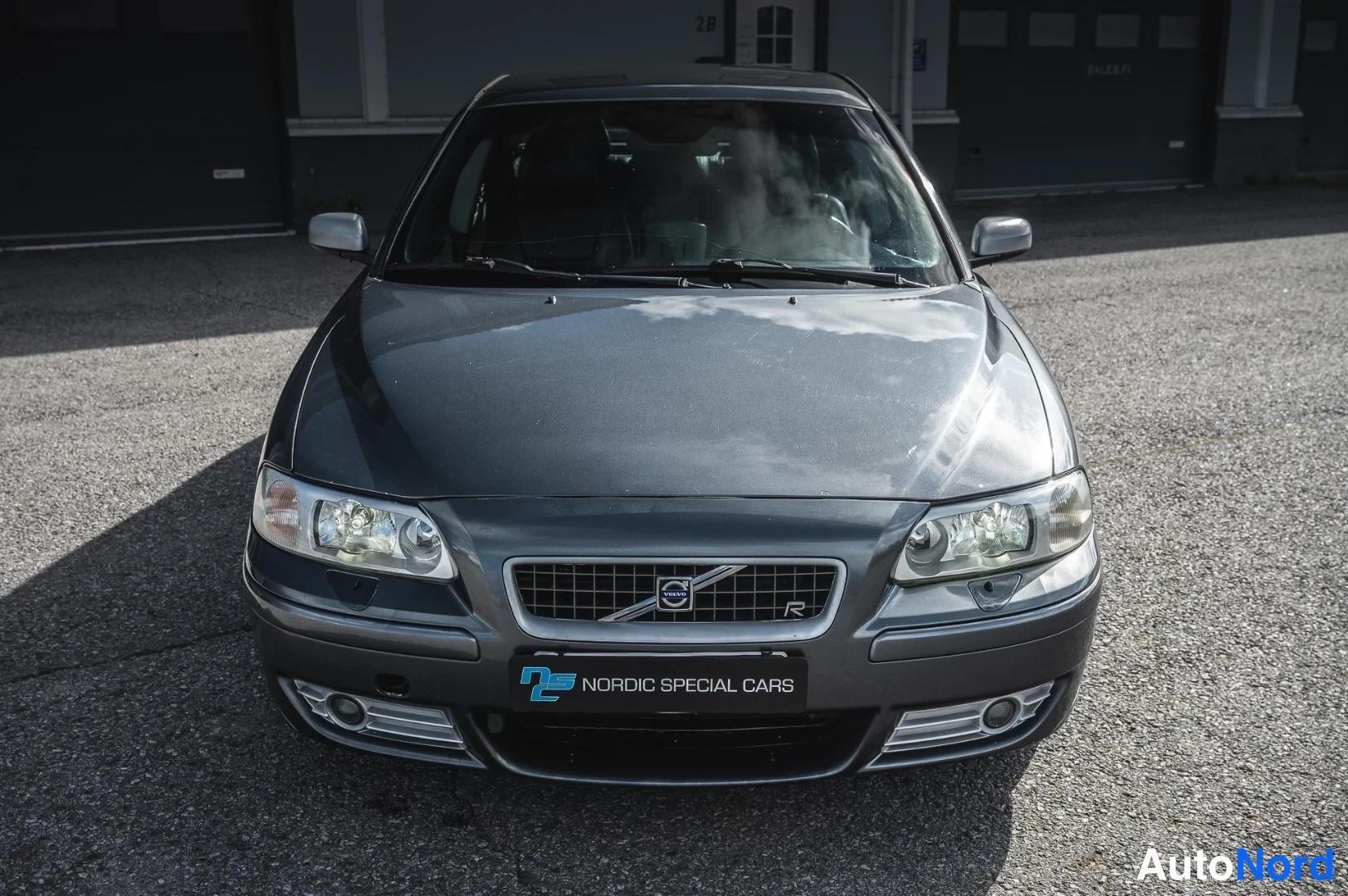Edestä: Auton suora etukuva: tummanharmaa S60, verkkokori Volvo-merkillä, sumuvalot ja Nordic Special Cars -kilpi.
