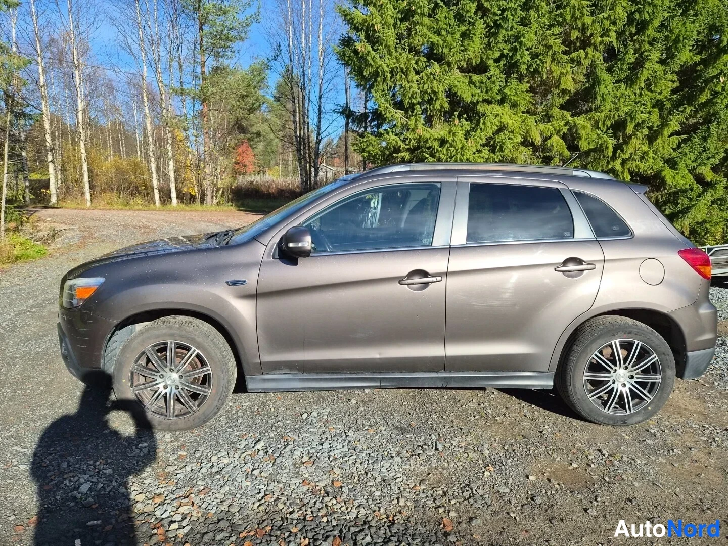 Sivulta: Ruskeanharmaa Mitsubishi ASX sivulta vasemmalta sorapohjaisella pihalla, kattokaiteet ja kompassin kaltainen merkki A-pillarissa.