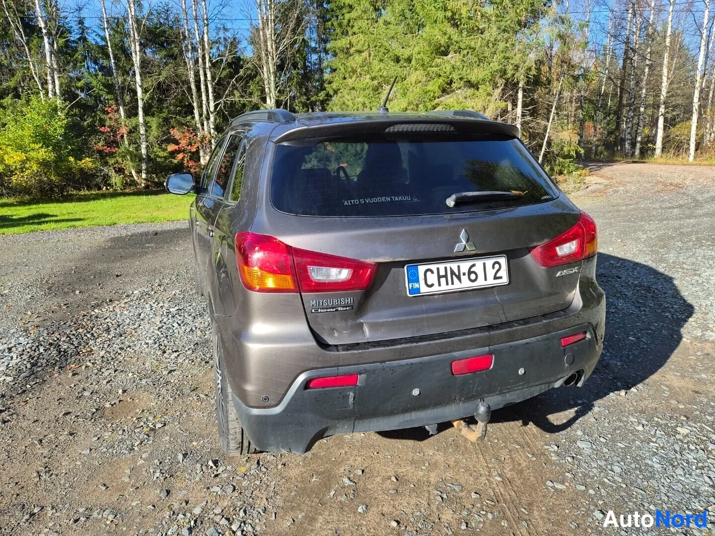Takaa: Auton perä suoraan takaa, Mitsubishi-merkki ja rekisterikilpi, sorapohjainen piha.