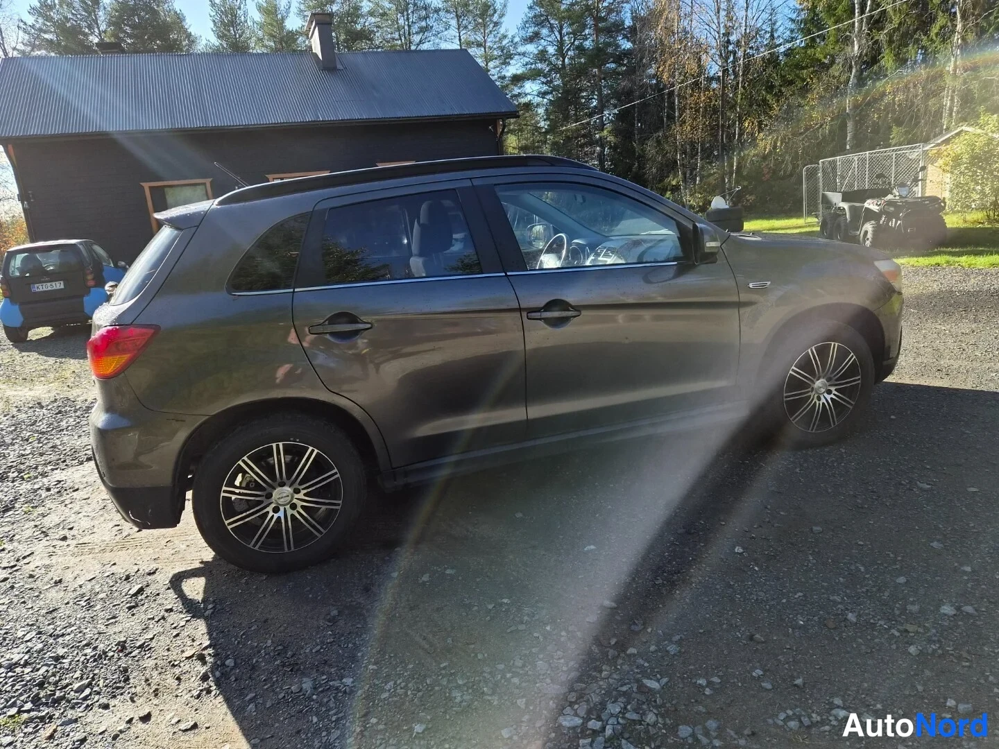 Takaviisto: Auton takakolmasosan sivukuva: tummanharmaa hatchback, takavalo ja kylki näkyvissä, autotalli ja metsä taustalla, kuva otettu sorapohjaisella pihalla.