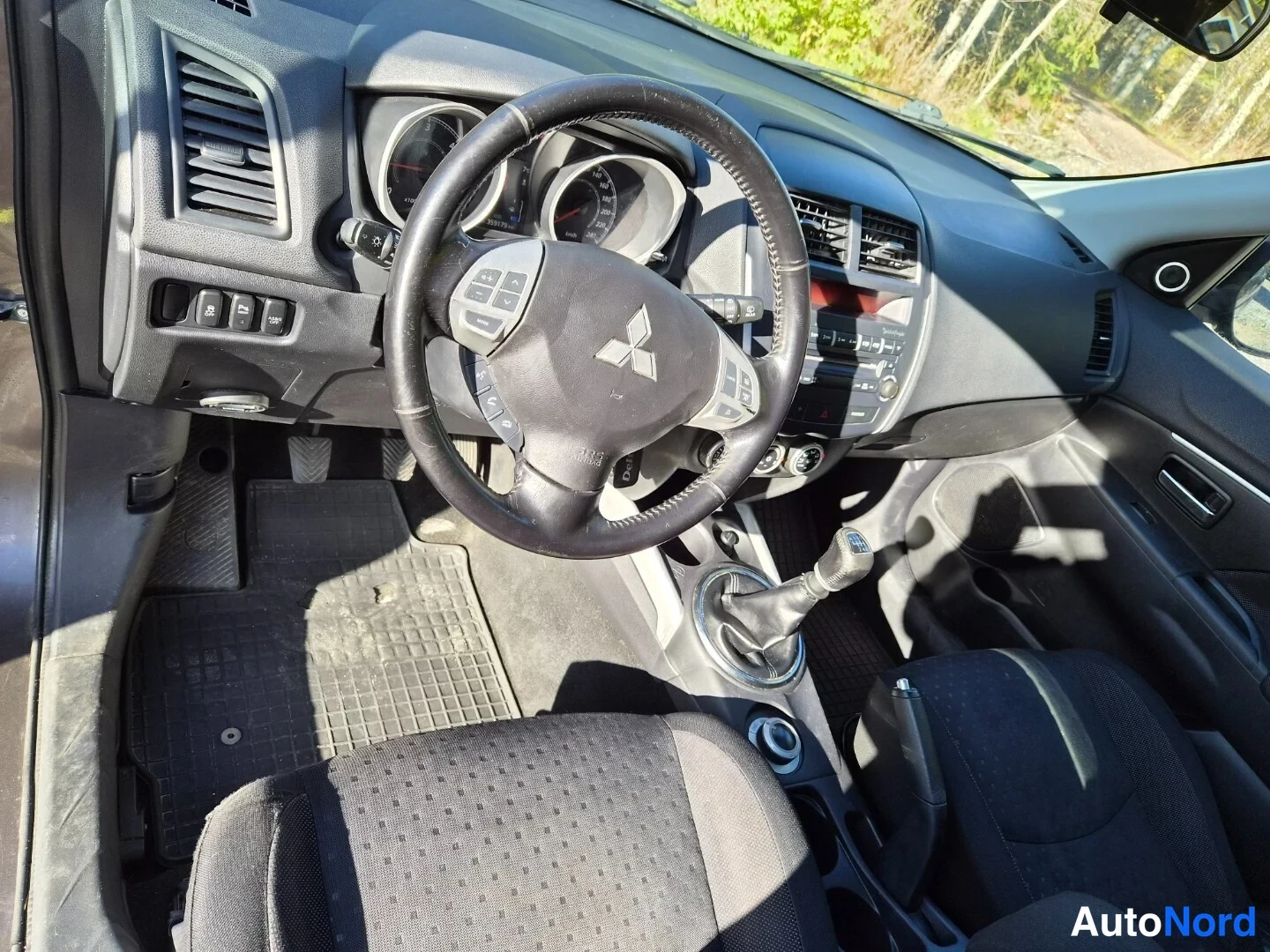 Edestä: Auton kuljettajan puolen sisätilat: ohjauspyörä Mitsubishi-tunnuksella, mittaristo, manuaalivaihteh circle ja tummat penkit.