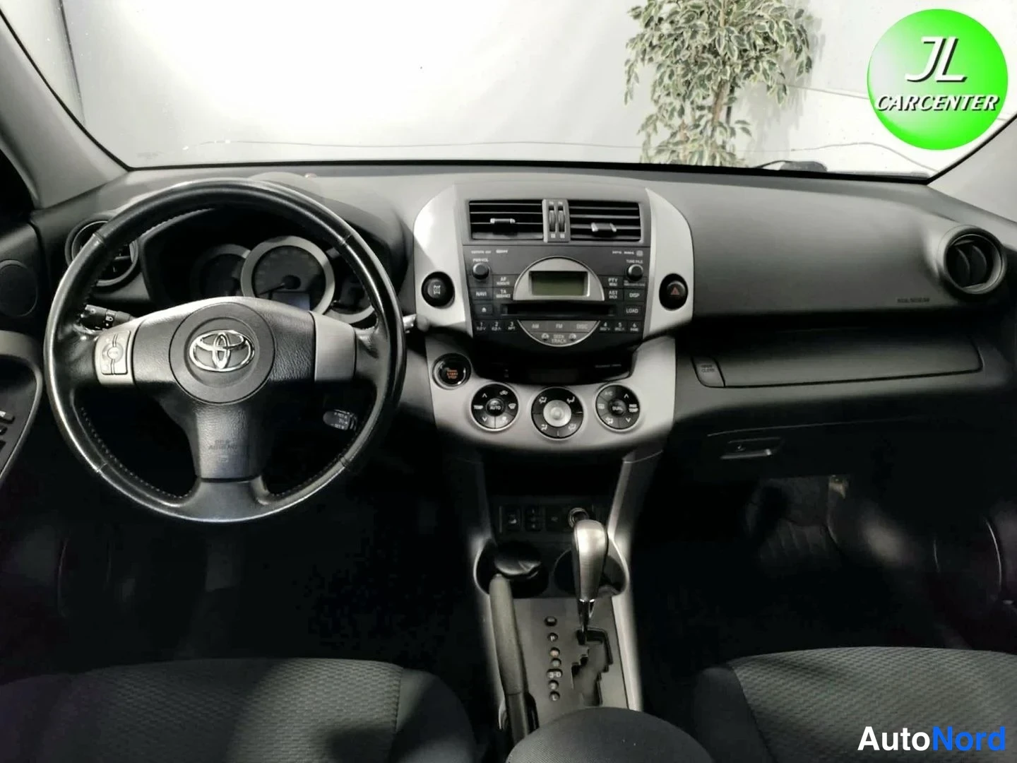 Muu: Auton kojetaulu ja keskikonsoli kuvattuna tuulilasin läpi: Toyota-logolla varustettu ohjauspyörä, radio ja vaihteenvalitsin.