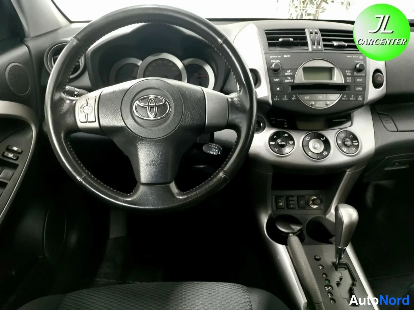 Muu: Lähikuva ohjauspyörästä ja keskikonsolista: Toyota-merkitty ohjauspyörä, radio- ja ilmastointilaitteet, automaattivaihteen valitsin.