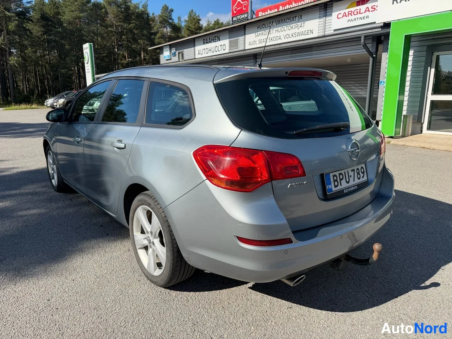 Takaviisto: Opel Astra -farmari takaovi ja takapää kuvattuina autokoripihan edessä, takavalo ja rekisteritunnus näkyvissä.