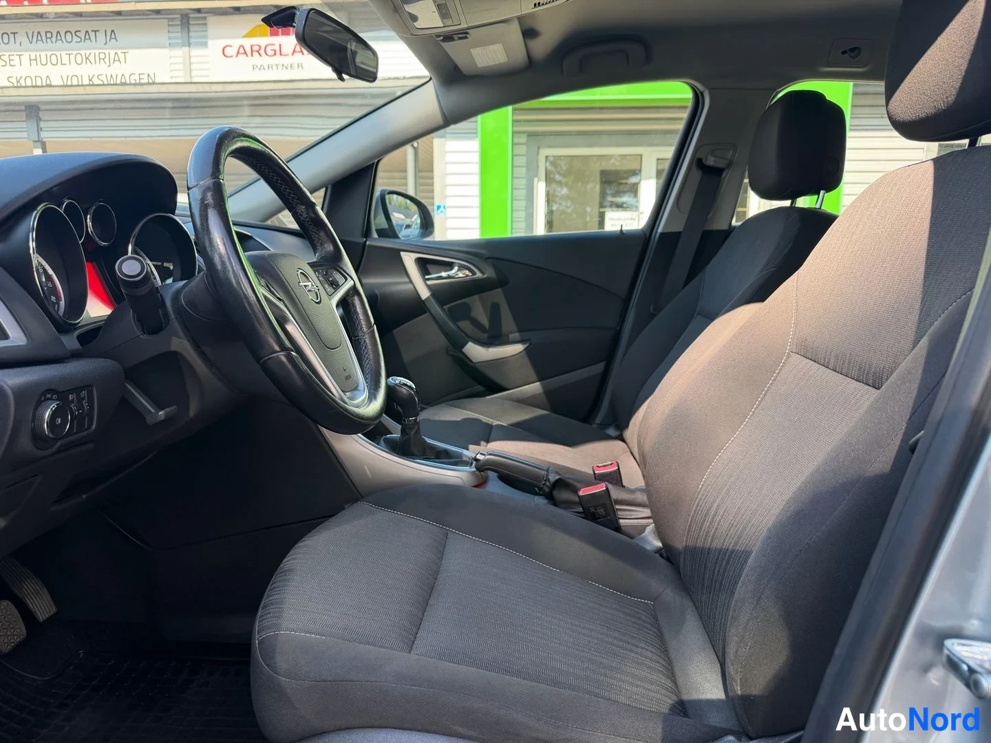 Muu: Ohjaamo sivulta: Opel-merkkinen ohjauspyörä, manuaalivaihdevipu, tummat kankaat istuimet ja instrumentit näkyvät.