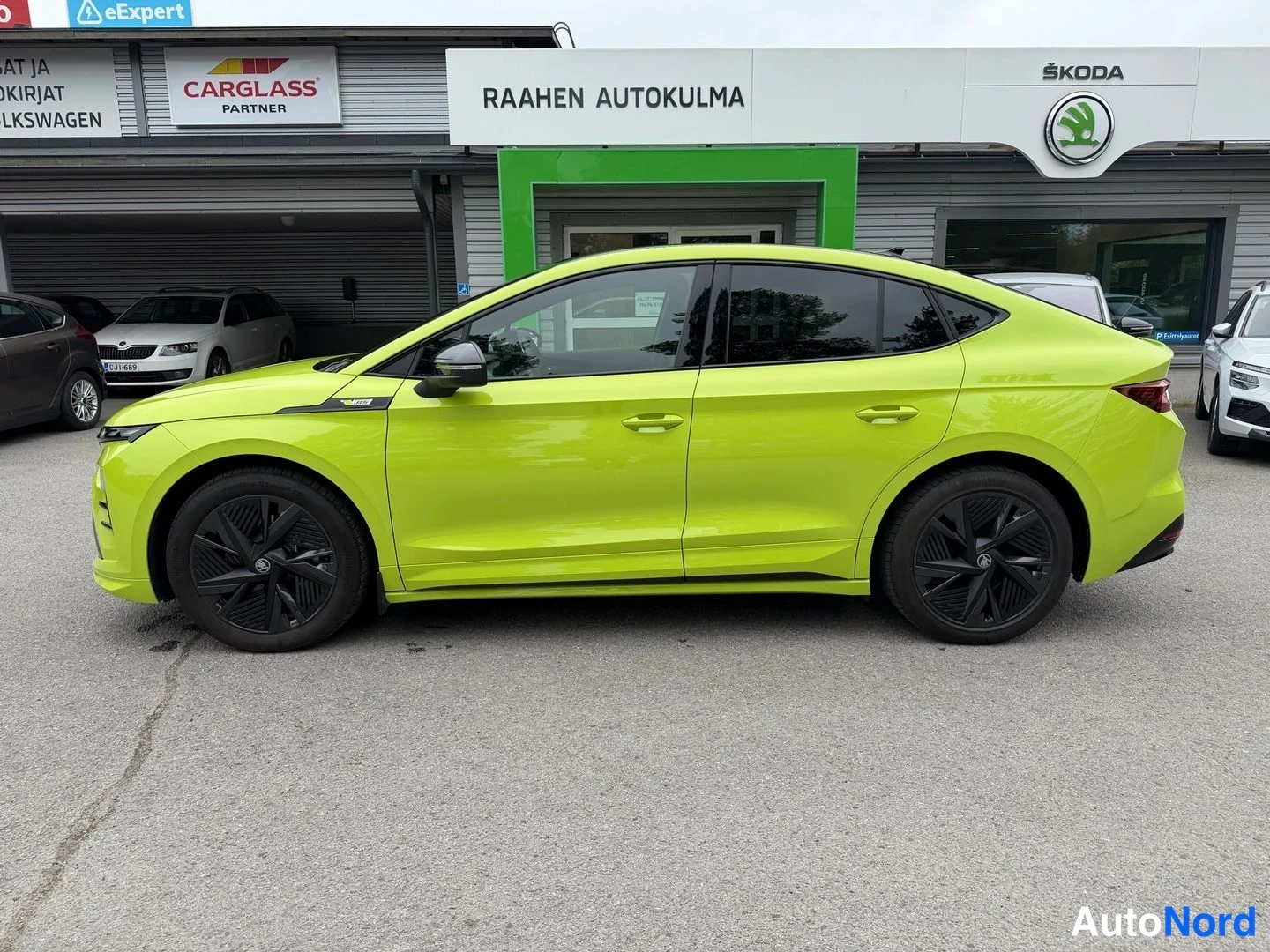 Sivulta: Kirkkaanvihreä Skoda Enyaq Coupe sivukuva autoluolan edessä, tummat vanteet ja tummat ikkunat.