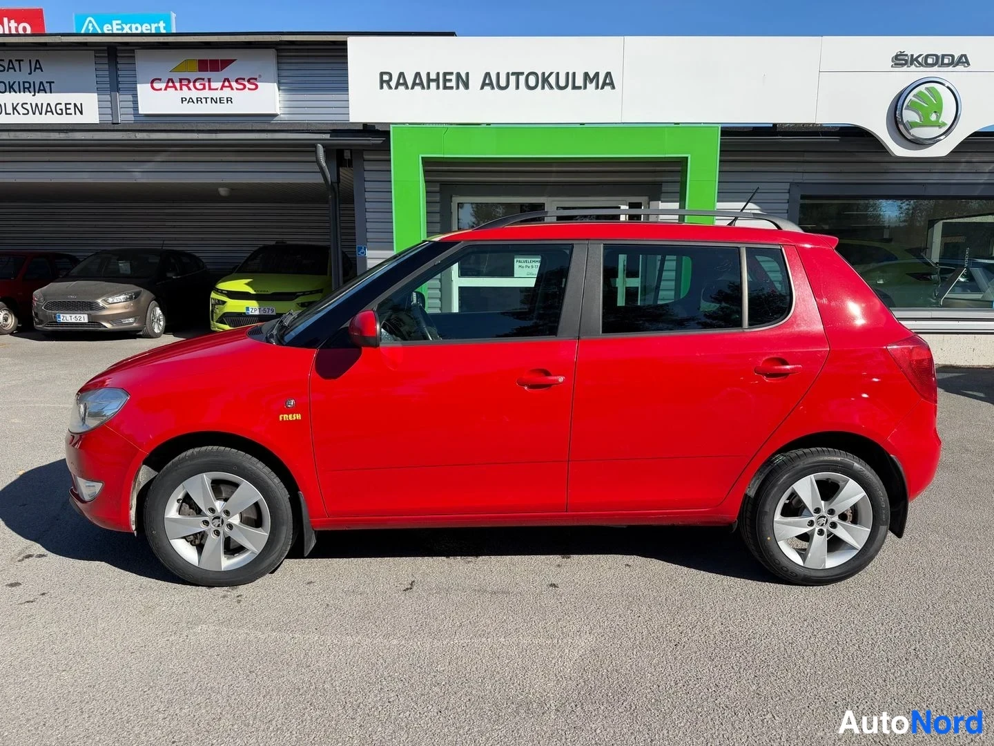 Sivulta: Punainen hatchback kylki- ja keulanäkymässä autoliikkeen edustalla, keula kuvan vasemmalla puolella.