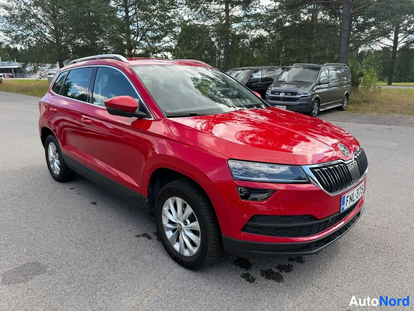 Etuviisto, nokka oikealle: Punaisen automobiilin etuosa kolmannelta kulmalta keulapuoli oikealla, rekisterikilpi, taustalla puita ja pakoautoja.