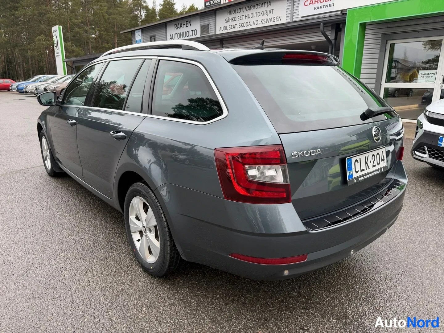 Takaviisto: Tummanharmaa farmariauto takaa vasemmalta kolmionneljanneskuva, ŠKODA-merkintä takaluukussa, rekisterikilpi ja punaiset takavalot näkyvät pihan parkkipaikalla.