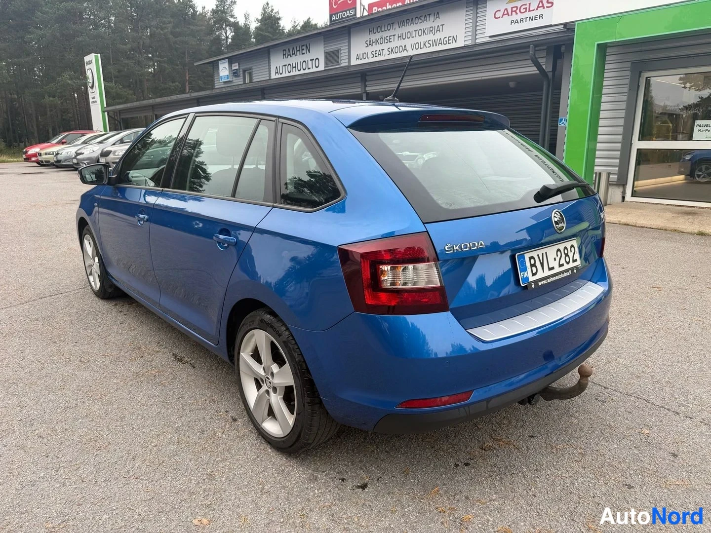 Takaviisto: Taas takaa vasemmalta kuvattu sininen Skoda Rapid Spaceback seisoo autotalon edustalla. Takavalo, rekisterinumero, vetokouku ja hopeiset vanteet ovat näkyvissä.