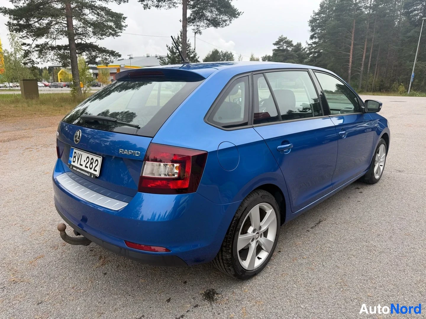 Takaviisto: Sininen Skoda Rapid -farmari takaviistosta nähtynä; rekisterikilpi, laajat takavalot, kattospoileri ja takaosan pieni lievä naarmu maalipinnassa.