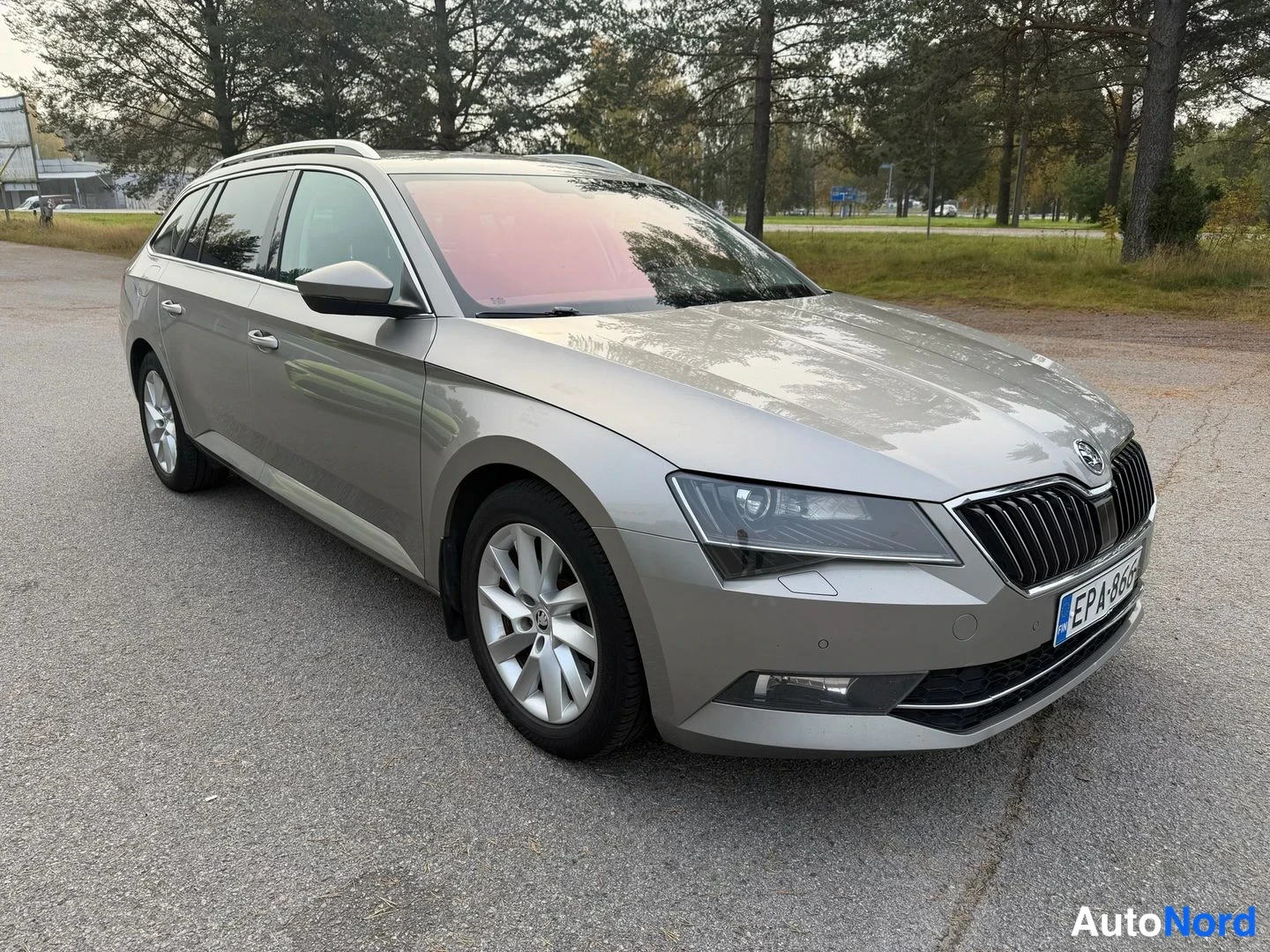 Etuviisto, nokka oikealle: Beige Kiina-auton Farmari parked on an asphalt lot with trees in the background, photographed from a front three-quarter angle showing the front face and