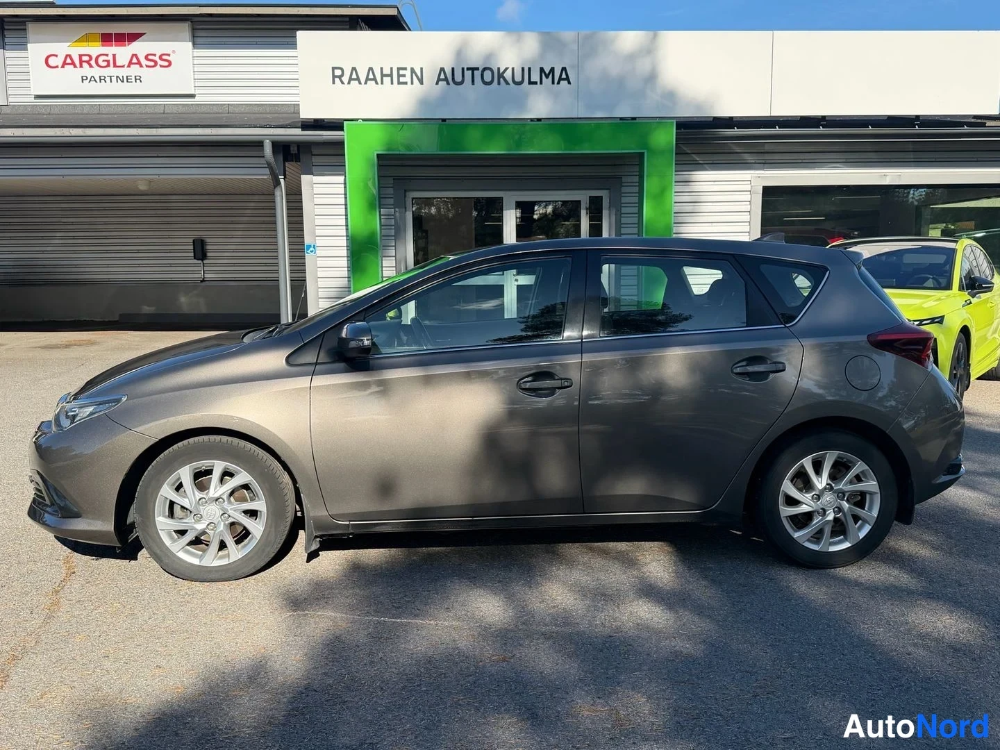 Etuviisto, nokka vasemmalle: Ruskeanharmaa Toyota Auris -henkilöauto sivusta vasemmasta etukulmasta autoliikkeen edustalla.