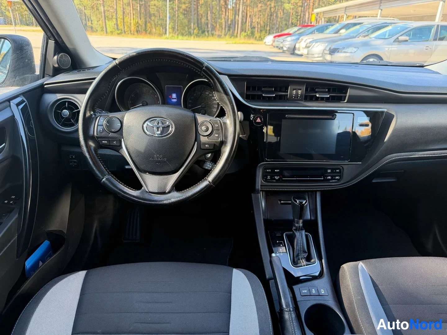 Etuviisto, nokka vasemmalle: Ohjaamo vasemmalta ohjatulla autolla: Toyota-merkkinen ratti, kojetaulu ja keskikonsoli, jossa automaattivaihteisto ja kosketusnäyttö.