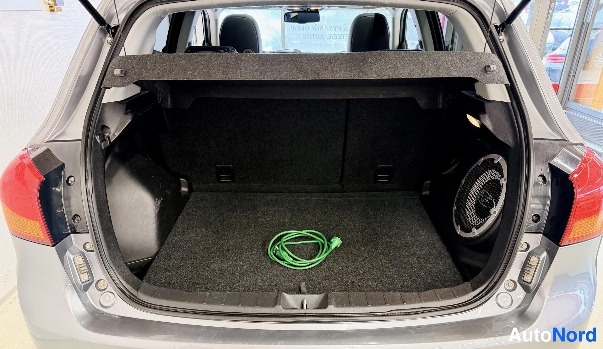 Takaa: Auton avattu tavaratila, musta matto ja vihreä kaapeli keskellä, subwoofer oikealla.