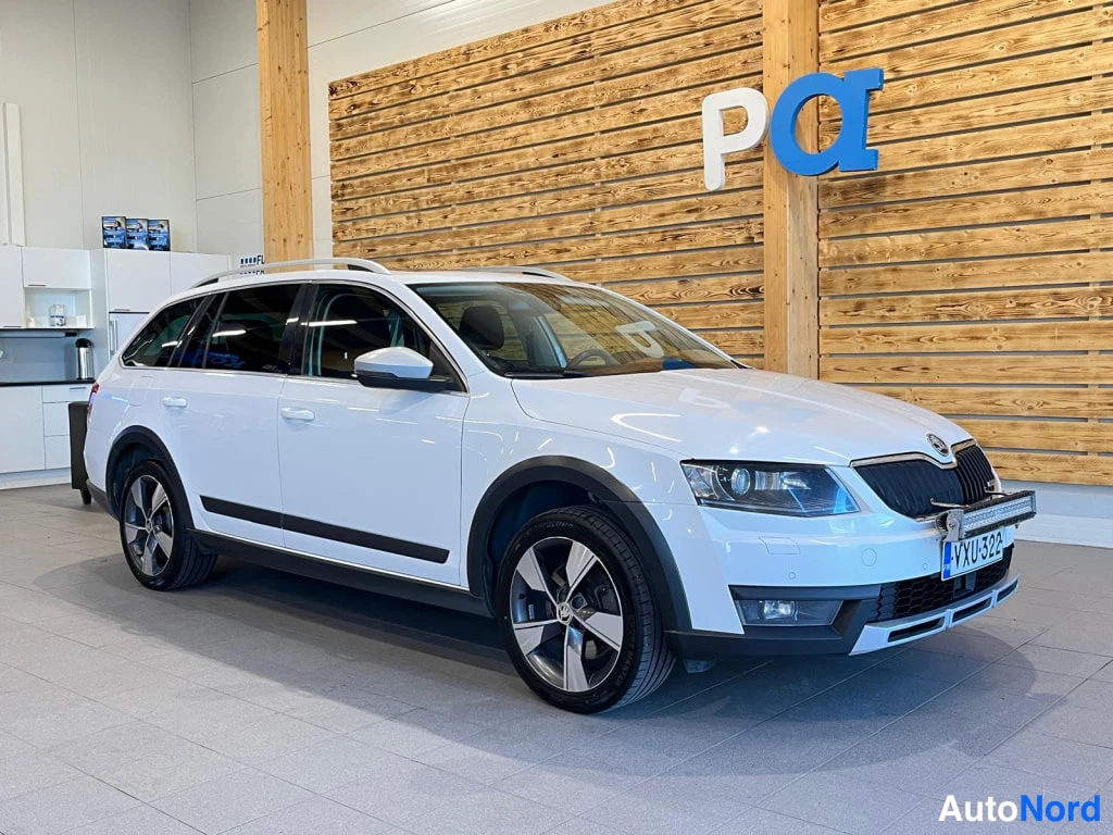 Etuviisto, nokka oikealle: Front three-quarter view showing the front grille, headlights, bonnet and left flank of a white station wagon parked inside a showroom.