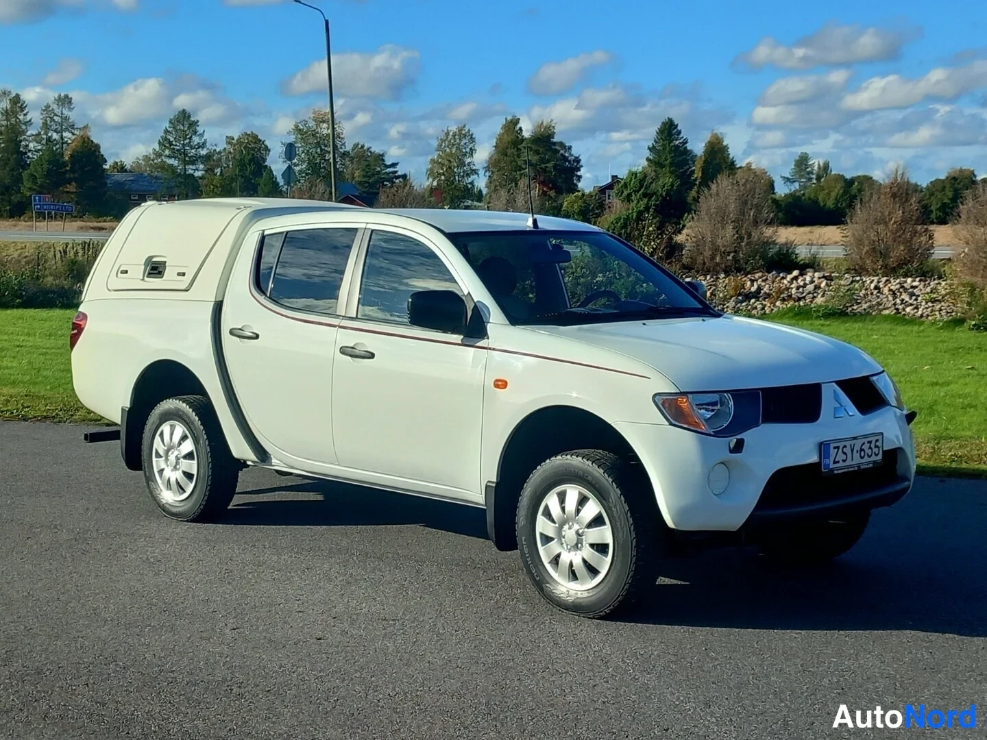 Etuviisto, nokka oikealle: Valkoinen Mitsubishi L200 -avolava etuviistosta oikealta, rekisteritunnus, neljä ovea ja lisäkori lavalla.