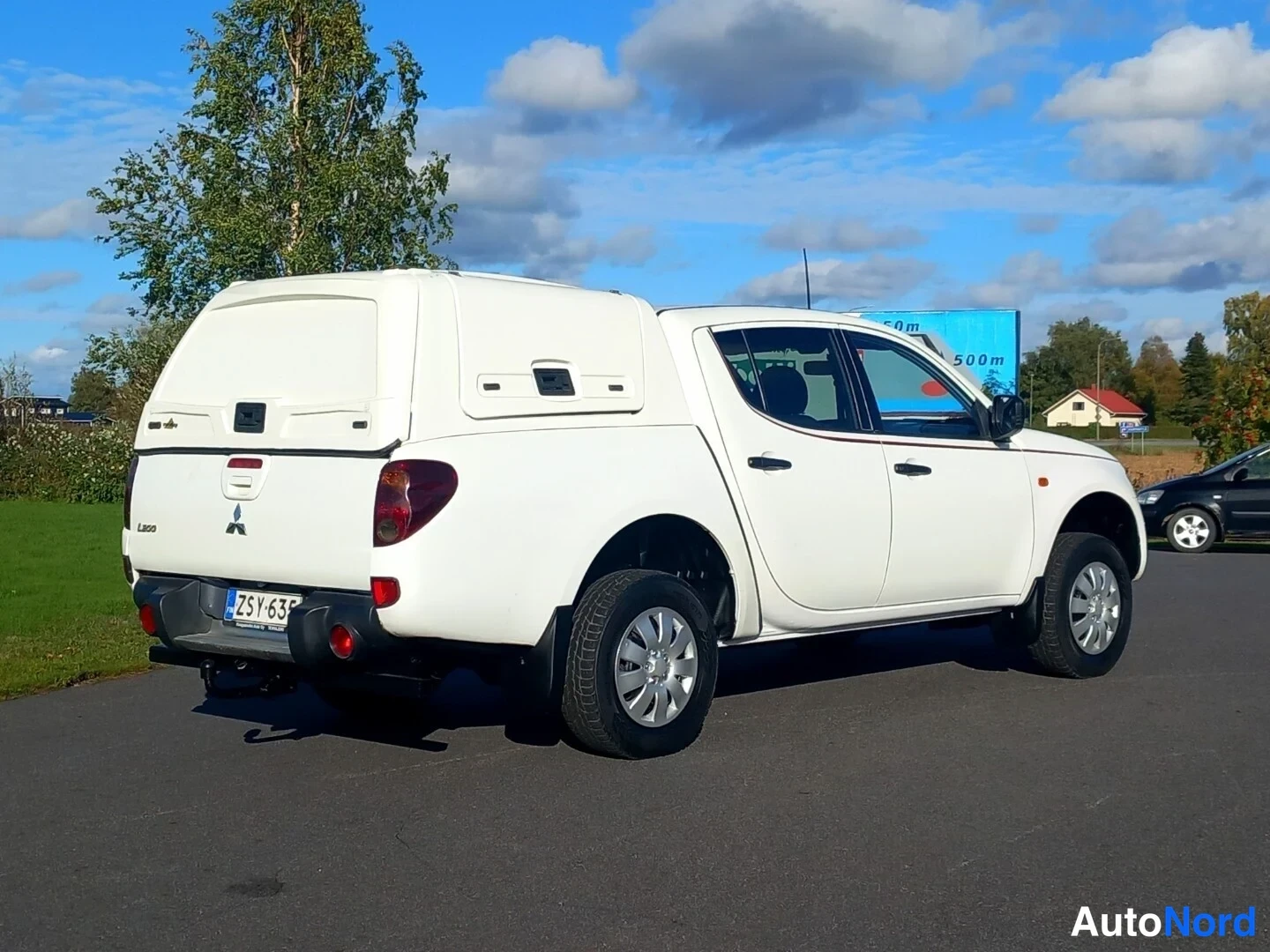 Takaviisto: Valkoinen Mitsubishi L200 takaviistosta vasemmalta, rekisteritunnus, lisäkori lavalla.